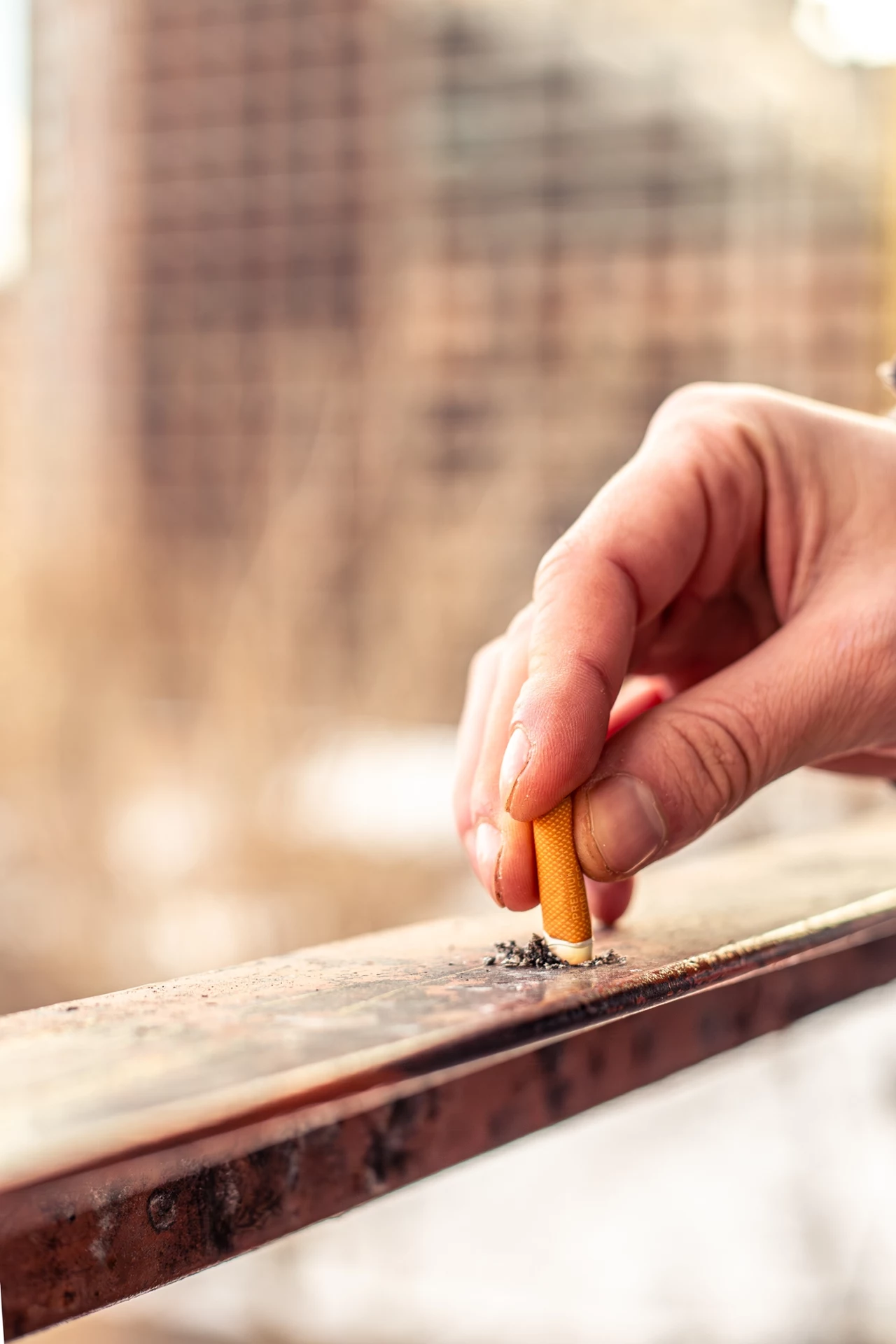 Palenie, kiedy zagraża innym sąsiadom, może podlegać karze. Dłoń gasząca papierosa na metalowej poręczy, wokół leżą popiół i resztki tytoniu, w tle rozmyte zabudowania miejskie.