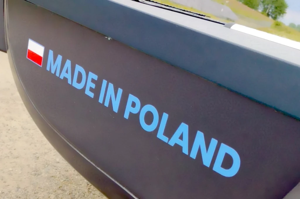 Firma z dumą podkreśla całkowicie polski rodowód samochodu Niebieski napis MADE IN POLAND oraz flaga Polski znajdują się na czarnym tle, fragment pojazdu, w tle widoczny asfalt i kawałek zieleni.