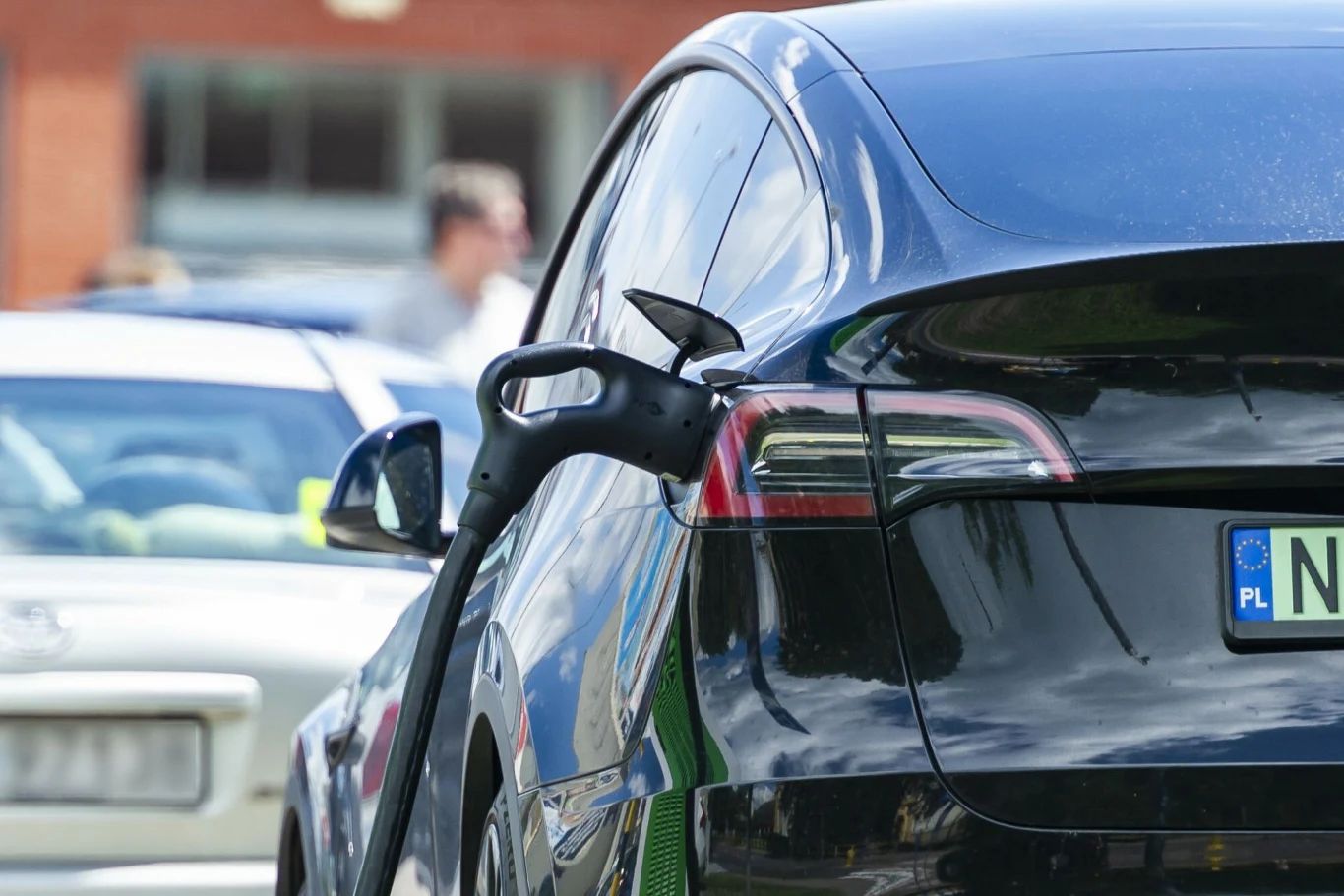 Program NaszEauto w liczbach. Nikt nie dostał jeszcze pieniędzy Elektryczny samochód podłączony do ładowarki na parkingu, w tle widoczne rozmyte sylwetki ludzi oraz inne pojazdy.