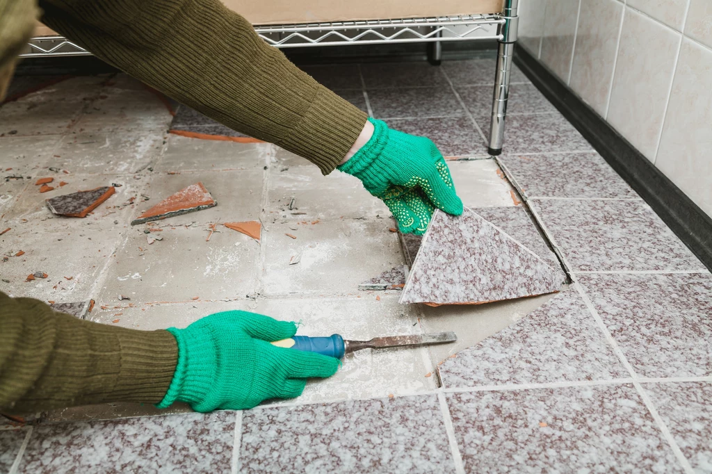 Ustal z sąsiadem, w jakich godzinach będzie wykonywać najcięższe prace remontowe Ręce w zielonych rękawicach podważają płytkę ceramiczną na podłodze za pomocą śrubokręta, w tle widać metalowy regał i resztki połamanych płytek.