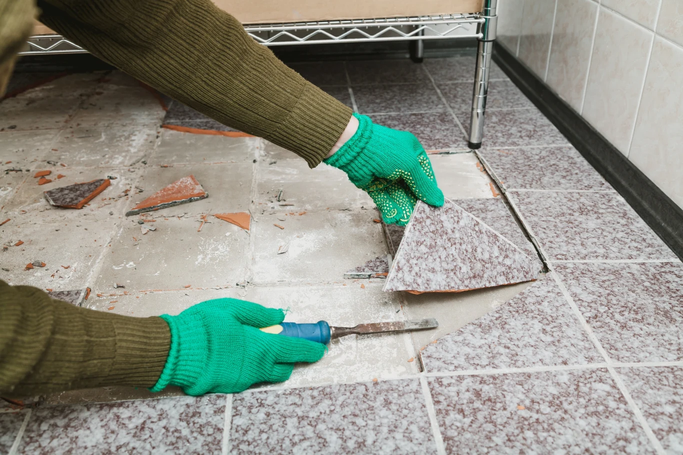 Ręce w zielonych rękawicach podważają płytkę ceramiczną na podłodze za pomocą śrubokręta, w tle widać metalowy regał i resztki połamanych płytek.