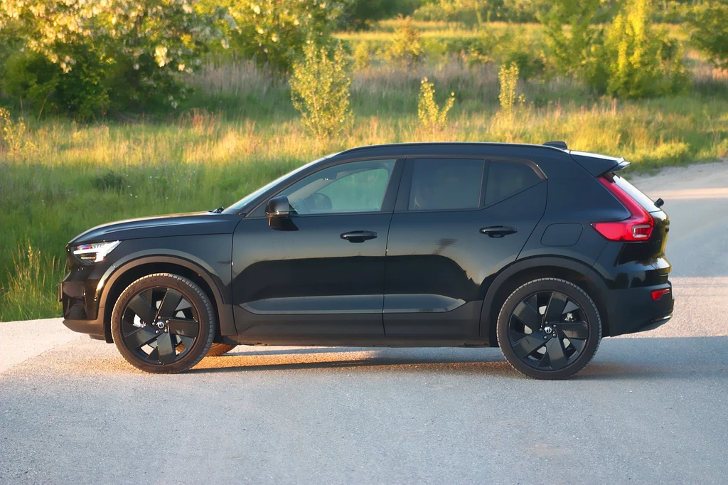 Volvo EX40 oparto na spalinowym modelu X40, doskonale znanym z polskich dróg Czarny samochód typu SUV stoi na pustej drodze asfaltowej w naturalnym, zielonym otoczeniu, na tle drzew i trawy oświetlonych zachodzącym słońcem.
