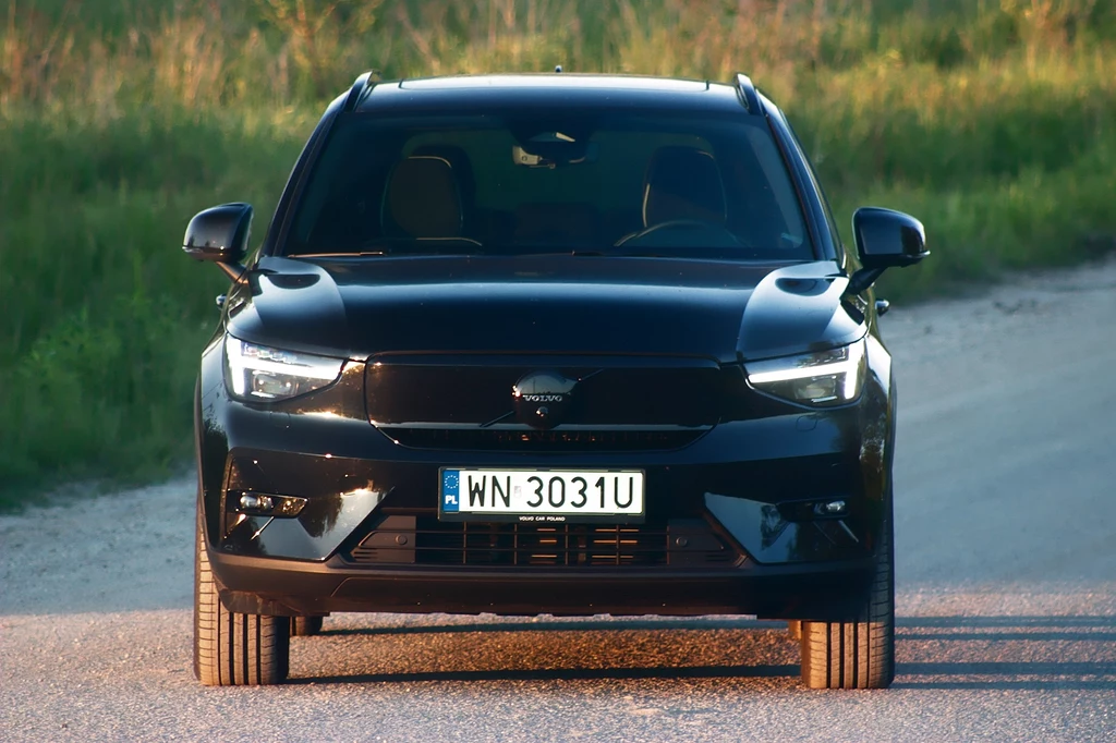 Wersja elektryczna do spalinowej różni się jedynie zabudowanym pasem przednim Czarny samochód marki Volvo stoi na wiejskiej drodze, widziany od frontu, z wyraźnie widoczną tablicą rejestracyjną oraz charakterystycznym logo producenta, otoczony zielenią w tle.