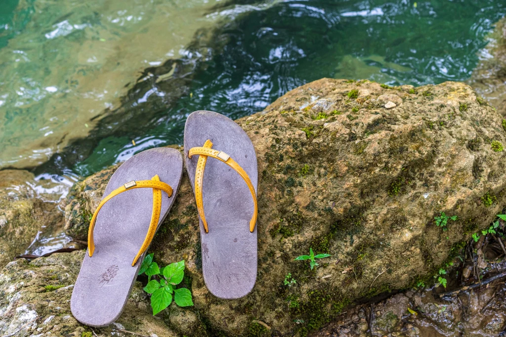 Najbardziej niezdrowe buty na lato Para szarych klapek z żółtymi paskami leży na omszałej skale obok czystej wody, wokół widoczne są zielone liście i naturalne otoczenie.