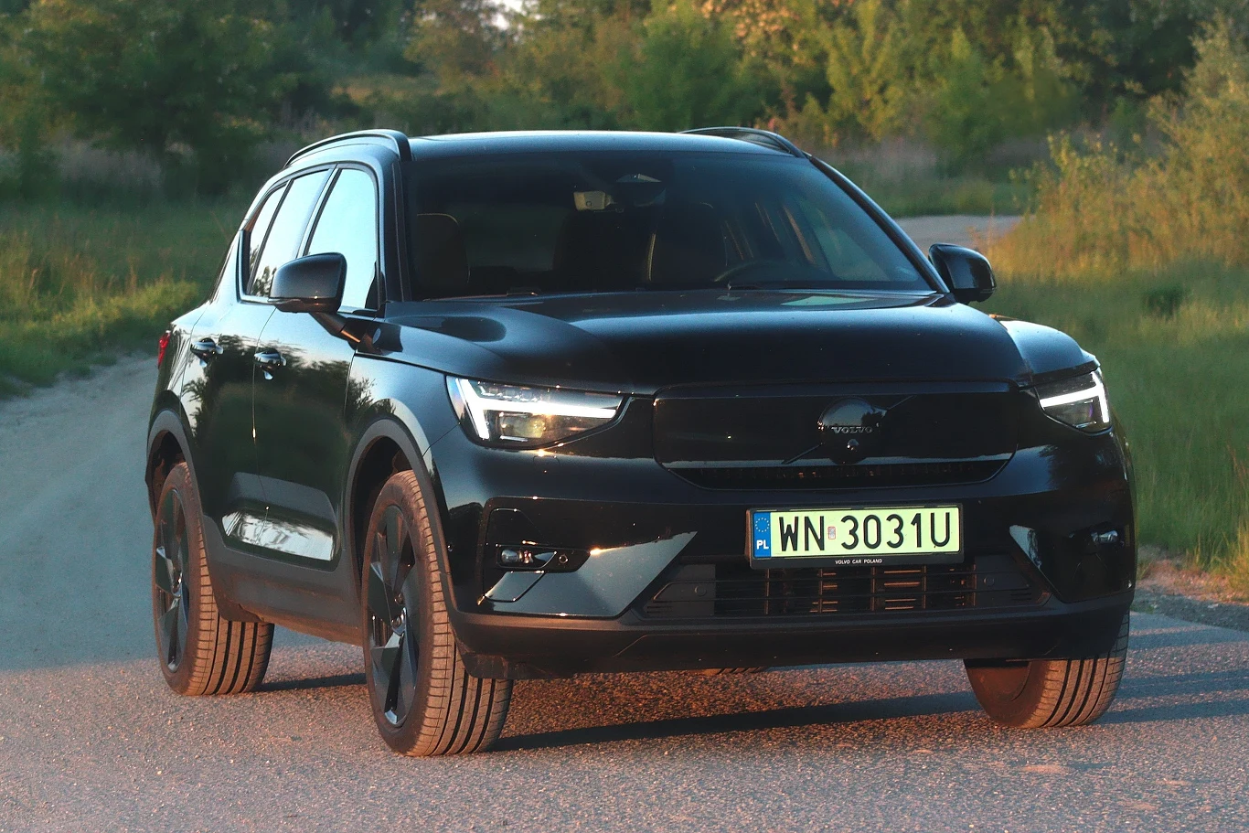 Czarny SUV marki Volvo stoi na żwirowej drodze, otoczony zielenią; samochód wyróżnia się nowoczesnym designem i reflektorami LED, na przednim zderzaku widoczna polska tablica rejestracyjna.