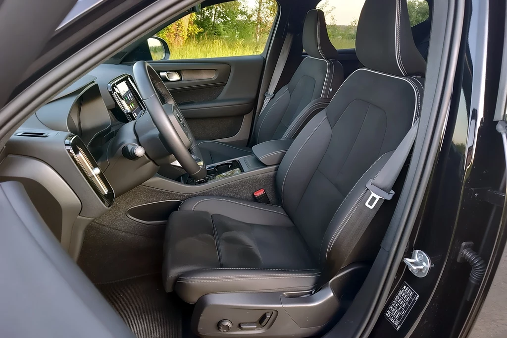 Fotele Volvo EX40 są bardzo wygodne Wnętrze nowoczesnego samochodu z czarnymi skórzanymi fotelami, deską rozdzielczą wyposażoną w ekran dotykowy i wielofunkcyjną kierownicą, przy otwartych drzwiach kierowcy, na tle zieleni widocznej przez okno.
