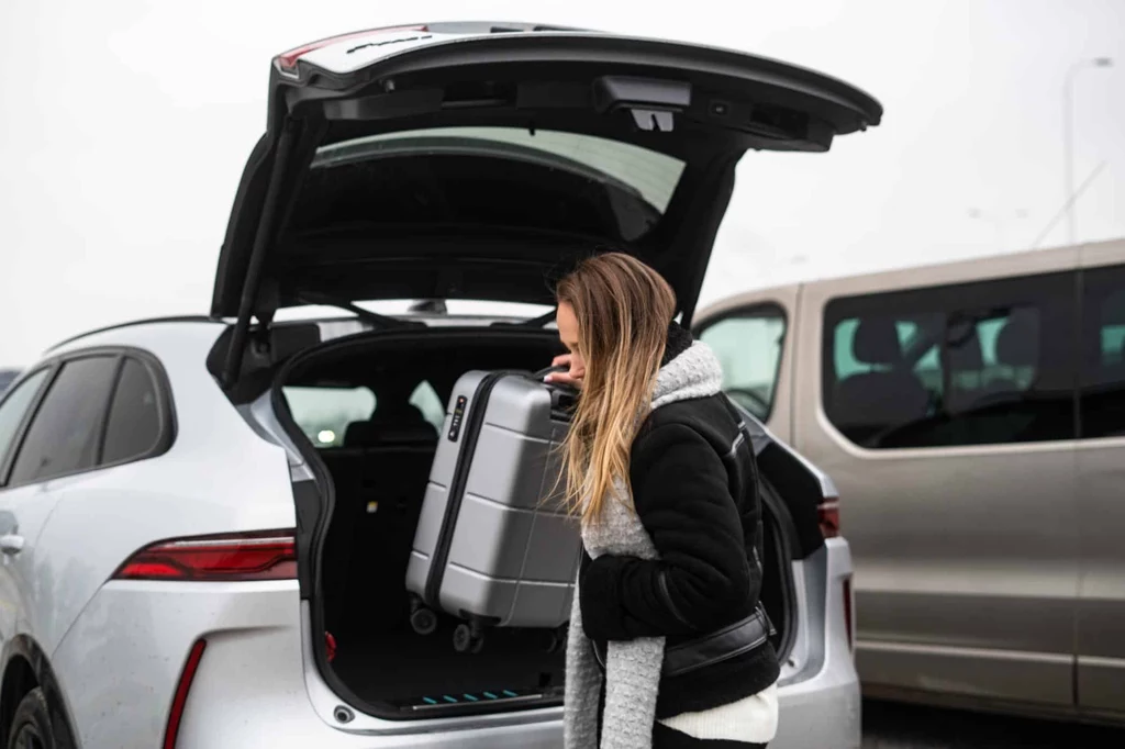 Parking: STANDARD, EXPRESS, SMART i VIP Młoda kobieta w ciepłych ubraniach wyciąga walizkę z bagażnika srebrnego samochodu osobowego, zaparkowanego na parkingu obok innego pojazdu.