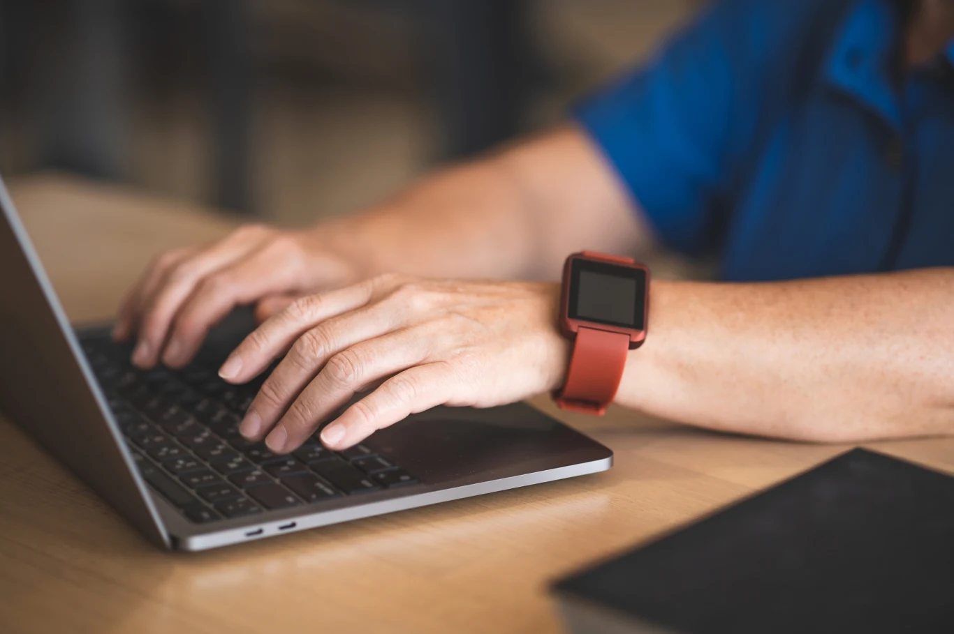 Smartwatch szpiegiem? Nawet komputer offline może wysyłać dane Osoba w niebieskiej koszuli korzystająca z laptopa, posiadająca na nadgarstku inteligentny zegarek z czerwoną opaską; na pierwszym planie fragment zamkniętego notesu lub książki.