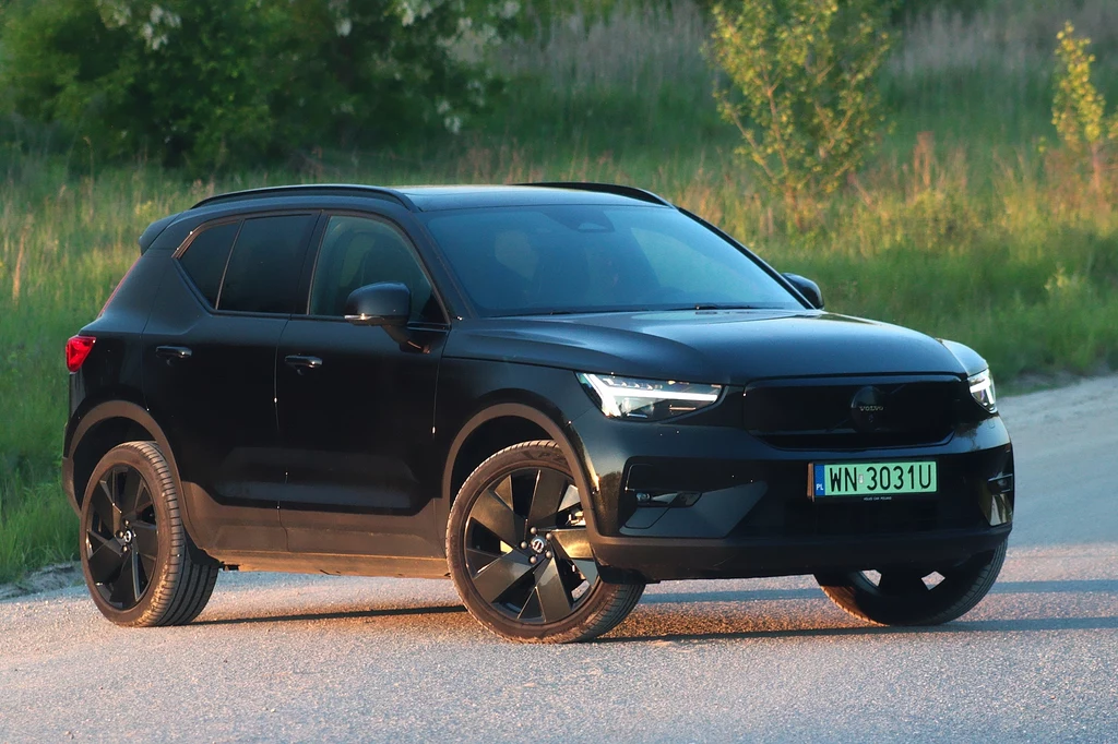 Volvo EX40 Czarny nowoczesny SUV zaparkowany na asfaltowej drodze, w tle zielona roślinność, auto prezentuje masywną sylwetkę i duże felgi, światła dzienne włączone, numer rejestracyjny widoczny z przodu pojazdu.