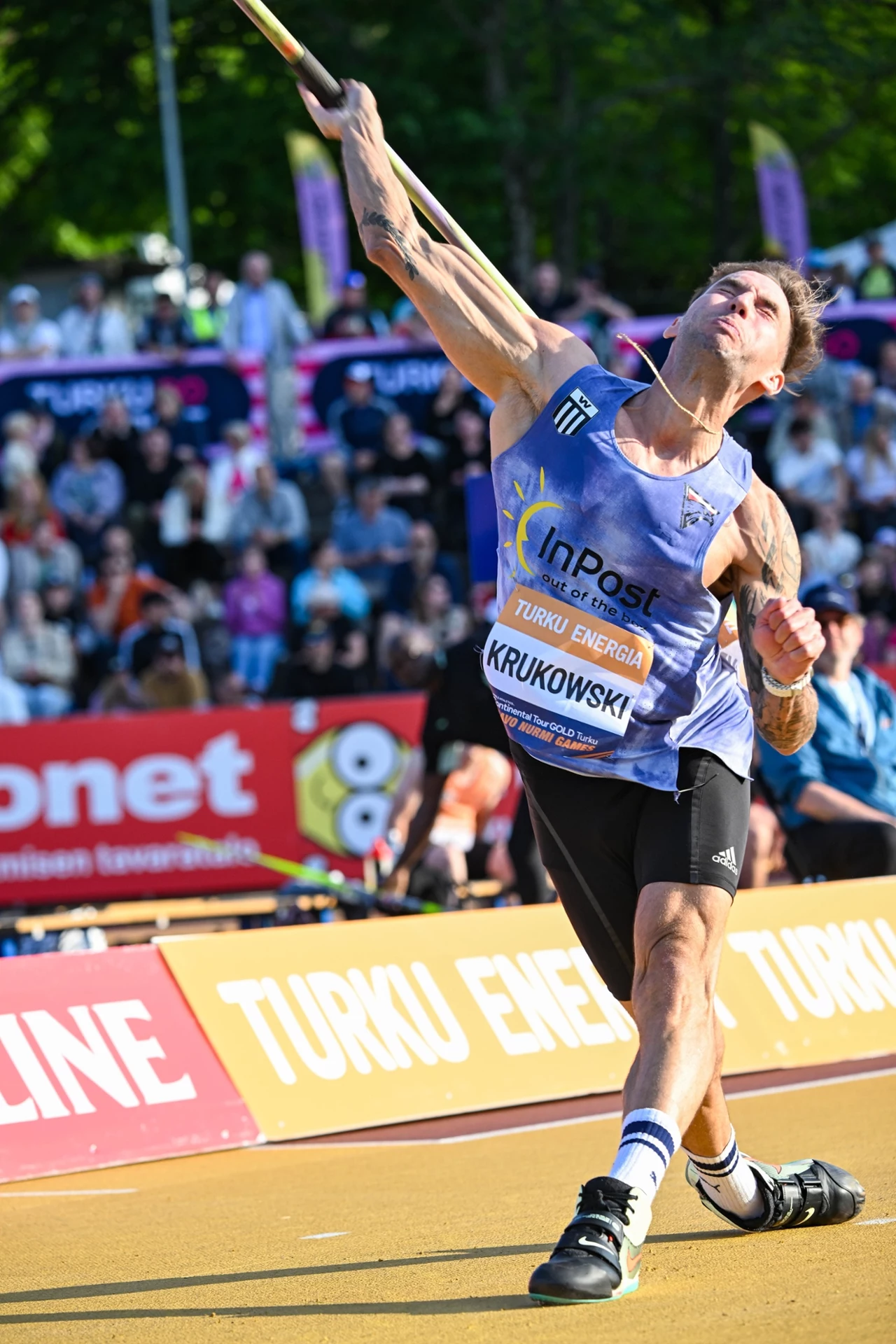 Marcin Krukowski Zawodnik w sportowej koszulce z numerem oraz nazwiskiem, wykonujący rzut oszczepem na stadionie podczas zawodów lekkoatletycznych; w tle widoczna zgromadzona publiczność i bannery reklamowe.