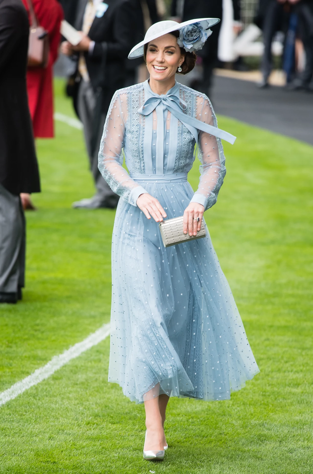 W 2019 roku księżna Kate zachwyciła błękitną, romantyczną kreacją na Royal Ascot Elegancka kobieta w długiej, jasnoniebieskiej sukni z przezroczystymi rękawami i zdobieniami, trzyma małą jasną kopertówkę, na głowie kapelusz ozdobiony dużym kwiatem, stoi na zielonej trawie, uśmiecha się, wokół osoby w eleganckich strojach.