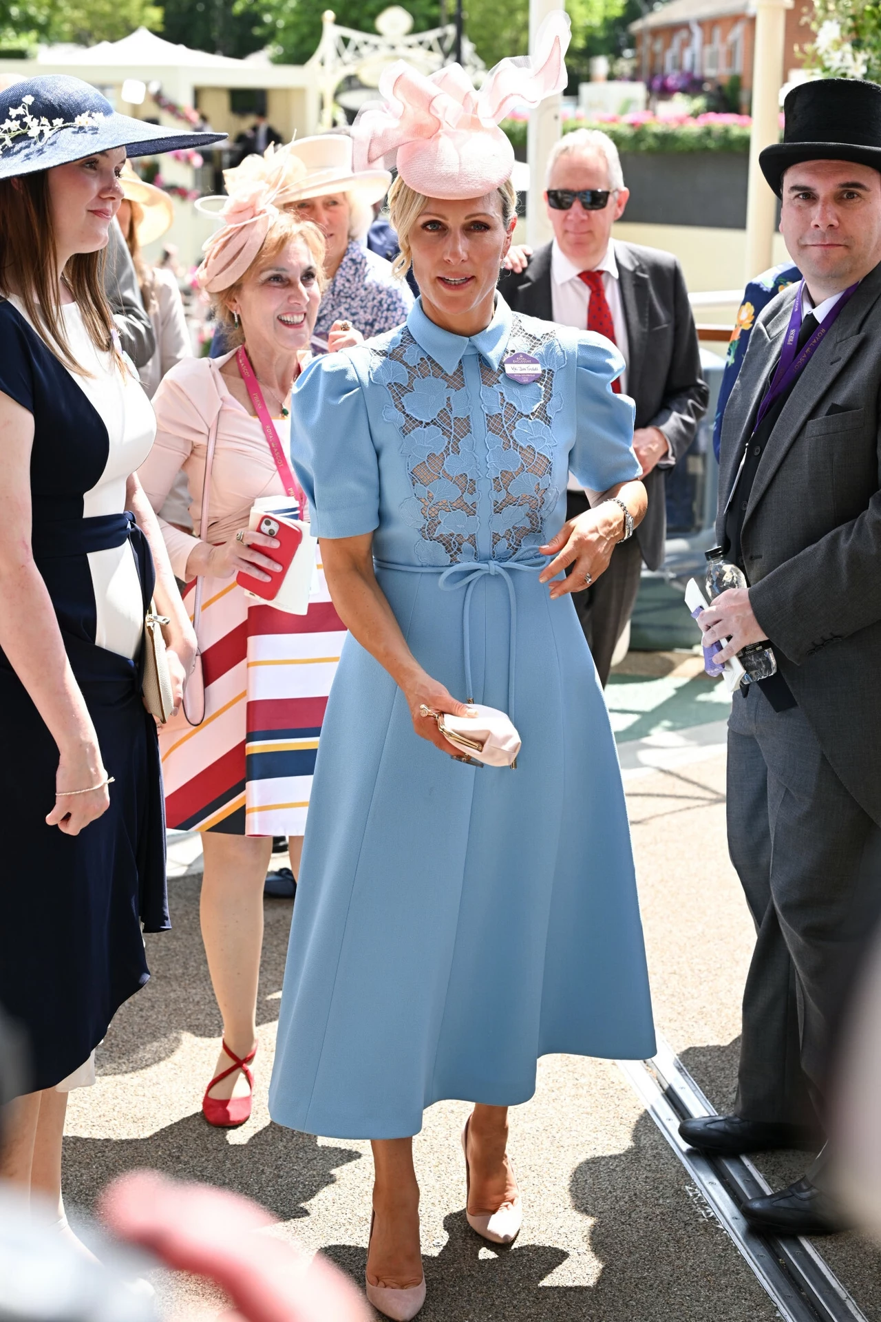 Zara Tindall nie nosi żadnego królewskiego tytułu. Tak postanowili jej ojciec i matka Elegancko ubrana kobieta w niebieskiej sukience z koronkową aplikacją i jasnoróżowym kapeluszem stoi w centrum grupy osób w letnich strojach i kapeluszach, w tle widoczne osoby rozmawiające i słoneczna pogoda.