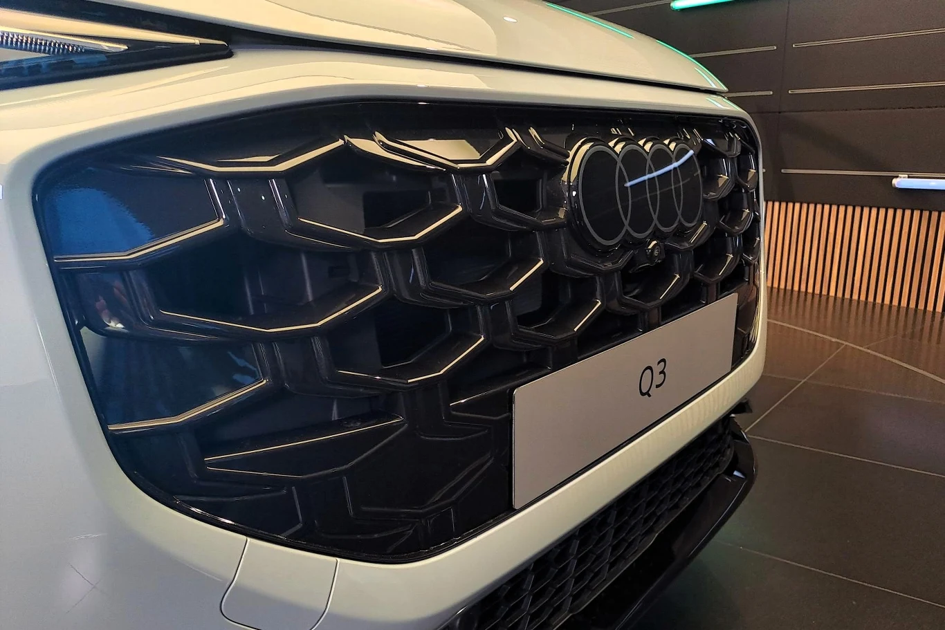 Duża, czarna, sześciokątna krata chłodnicy samochodu Audi Q3 z widocznym charakterystycznym logo marki Audi pośrodku oraz fragmentem białej karoserii, w tle nowoczesne wnętrze salonu samochodowego.