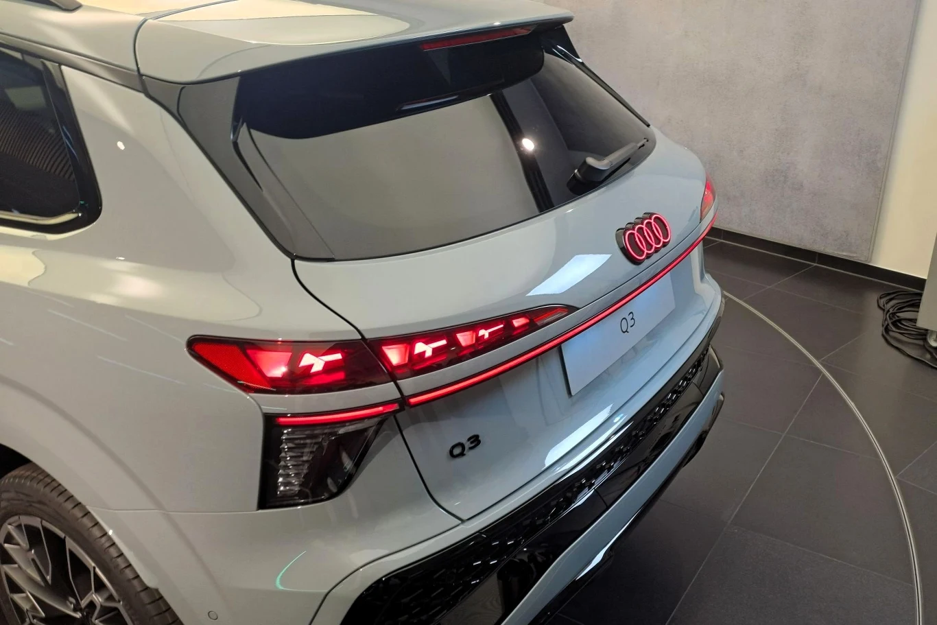 Tylna część srebrnego samochodu marki Audi Q3 z wyraźnie widocznymi nowoczesnymi lampami LED, chromowanym elementem na zderzaku oraz logotypem Audi na klapie bagażnika, pojazd ustawiony w jasnym, zamkniętym pomieszczeniu na czarnej posadzce