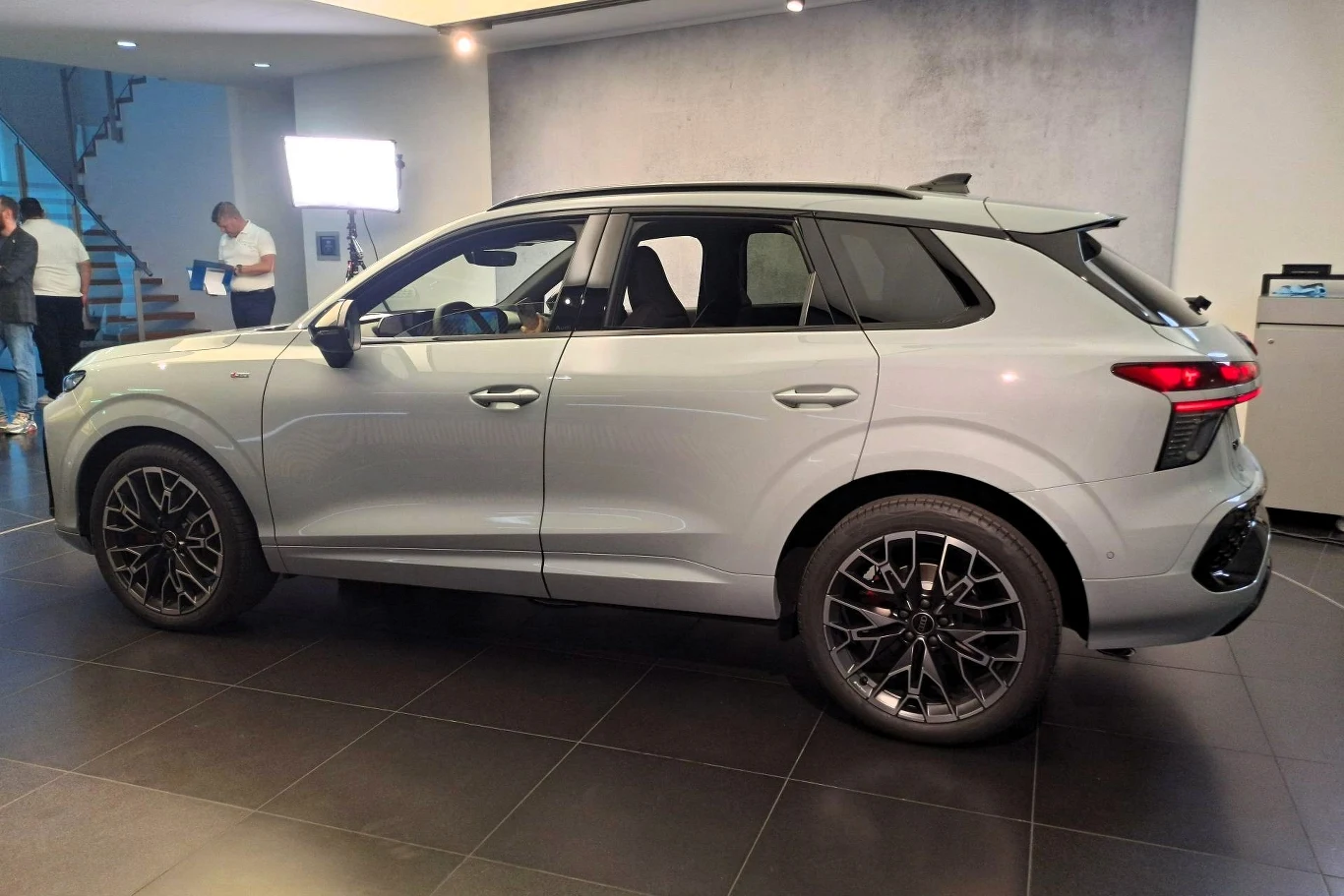 Srebrny samochód osobowy typu SUV stoi w nowoczesnym, jasnym wnętrzu na czarnej, połyskującej posadzce, w tle widoczne są osoby rozmawiające obok schodów oraz źródło światła do oświetlenia pojazdu.