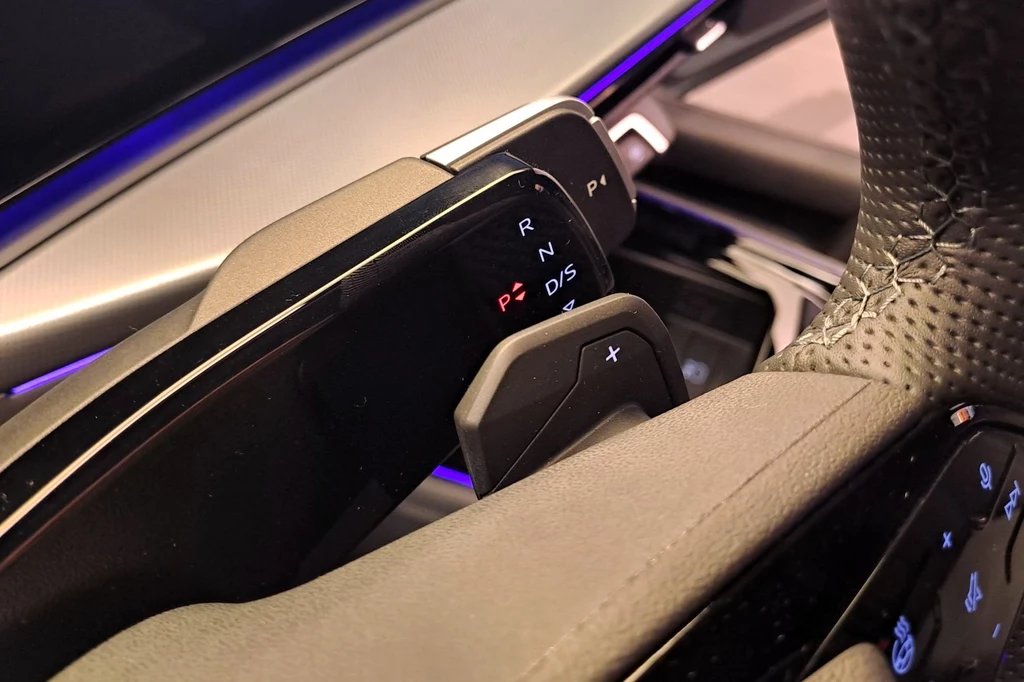 W nowym Audi Q3 dźwignia zmiany biegów została przeniesiona na kolumnę kierownicy i znajduje się po jej prawej stronie. Dźwignia po lewej integruje obsługę świateł i wycieraczek. Zbliżenie na panel zmiany biegów oraz fragment kierownicy nowoczesnego samochodu, podświetlenie LED w tle i widoczne przyciski sterujące biegiem i napędem.