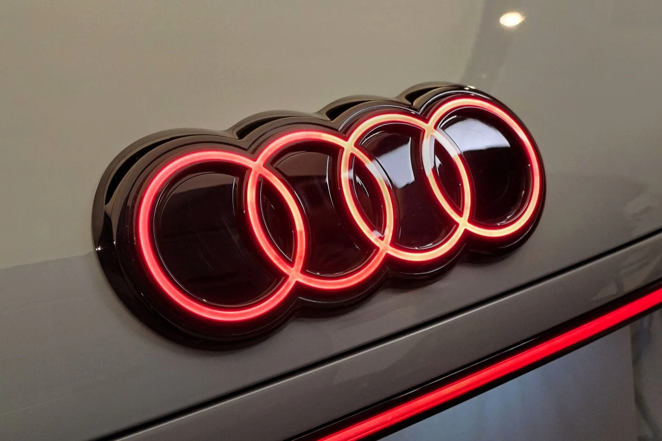 Podświetlone, czerwone logo Audi na błyszczącej powierzchni samochodu, z odbiciem światła na emblemacie.