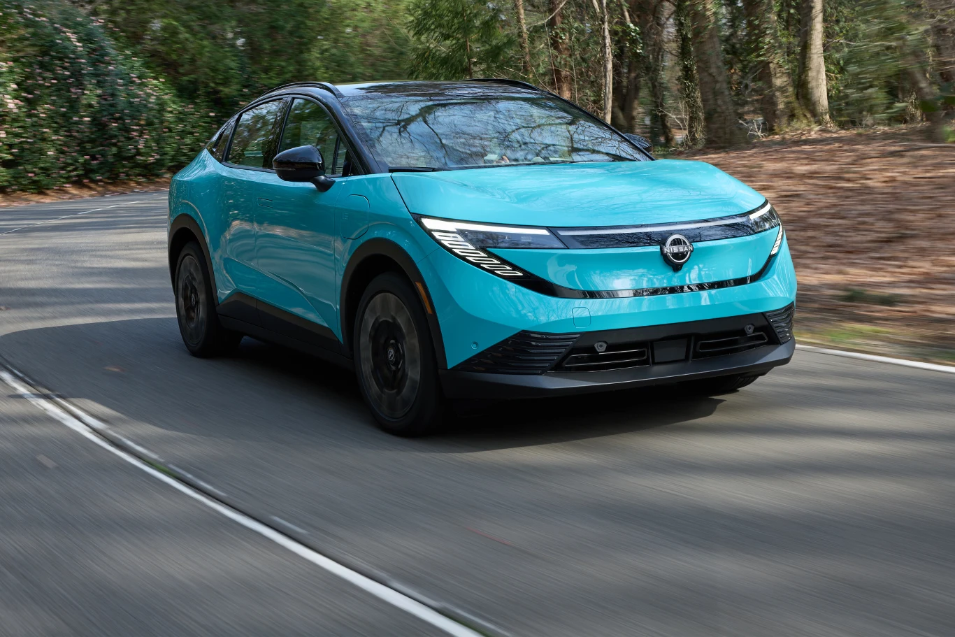 Nadjeżdża nowy Nissan Leaf. Pierwsze egzemplarze trafią do salonów wiosną przyszłego roku. Jasnoniebieski samochód elektryczny jedzie po asfaltowej, krętej drodze otoczonej zielonymi drzewami, prezentując nowoczesny design i aerodynamiczny kształt nadwozia.