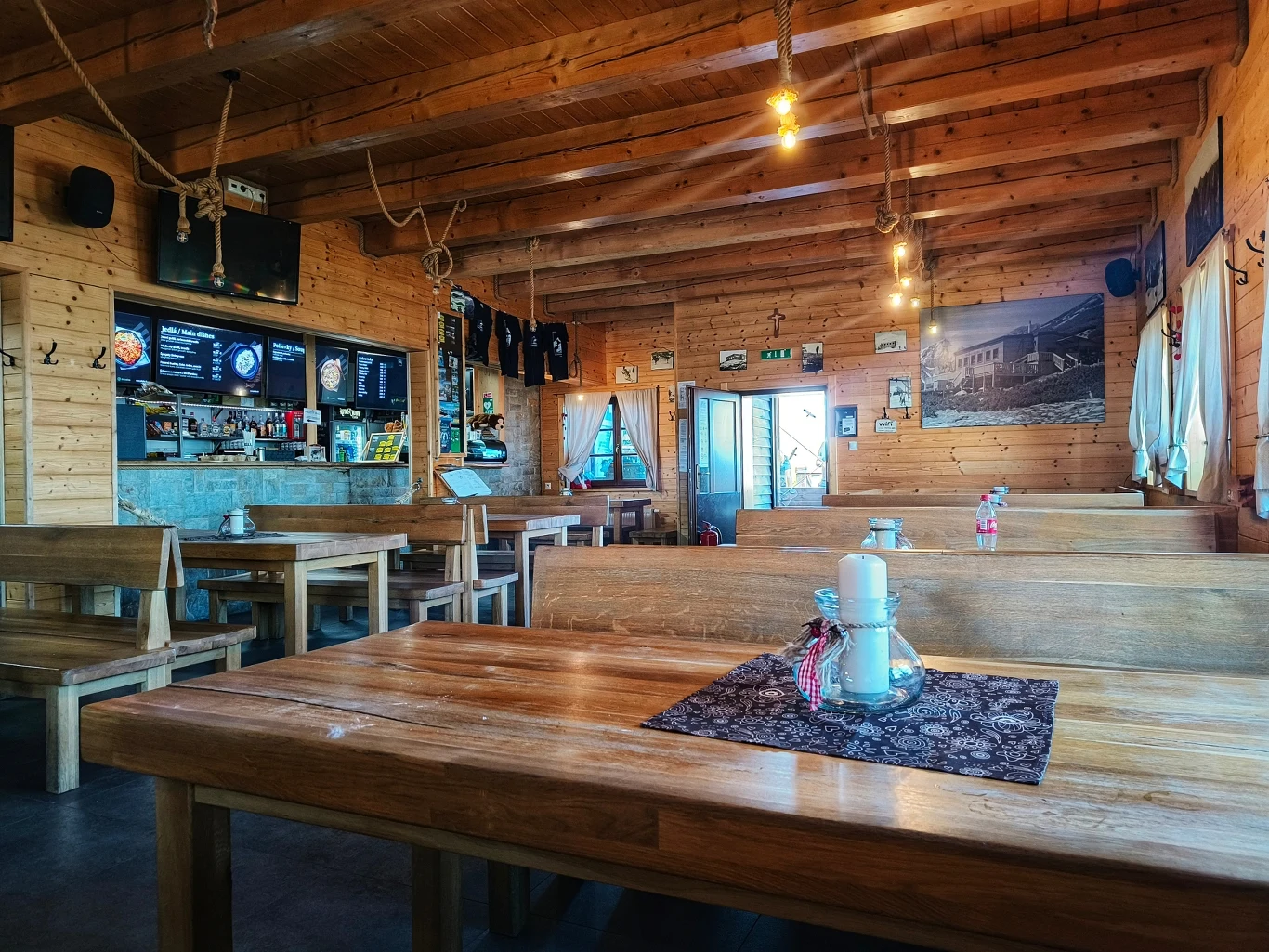 Drewniane wnętrze restauracji z ławami i stołami, ozdobnymi lampionami na środku stołów, jasnym światłem i widoczną ladą barową w tle, na ścianach znajdują się zdjęcia i pamiątki.