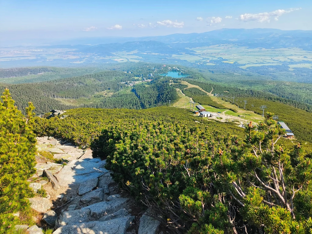 Nabieramy wysokości Kamienista ścieżka prowadząca przez gęste zielone krzewy pośród gór, w oddali rozpościera się rozległa panorama doliny z licznymi polami i lasami, widoczne są pojedyncze zabudowania na stokach.