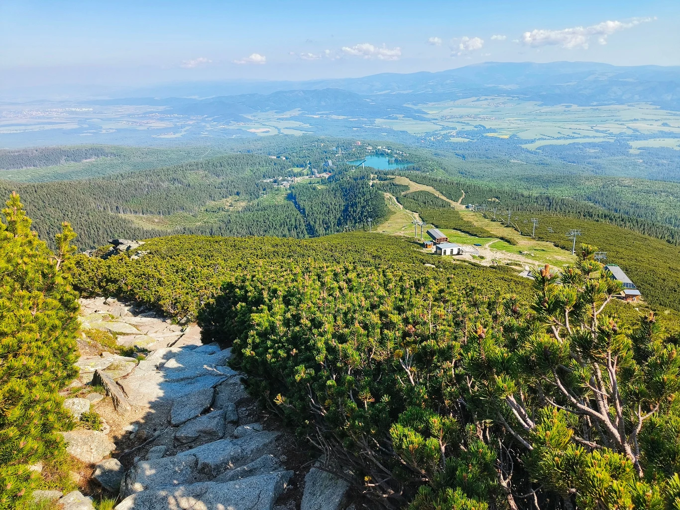 Kamienista ścieżka prowadząca przez gęste zielone krzewy pośród gór, w oddali rozpościera się rozległa panorama doliny z licznymi polami i lasami, widoczne są pojedyncze zabudowania na stokach.