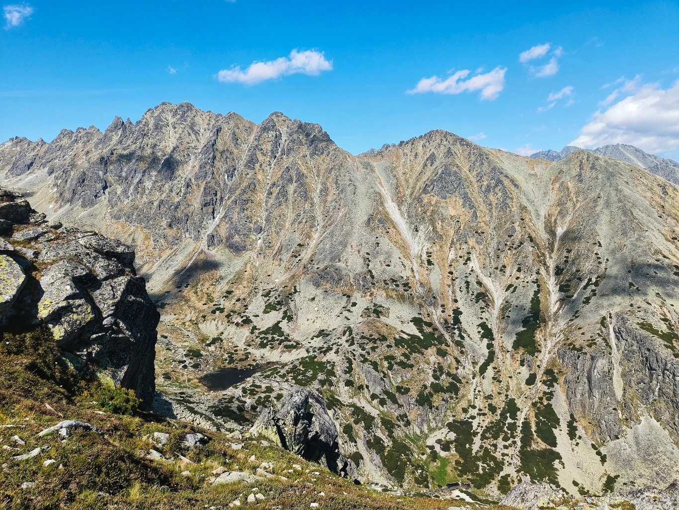 Stroma grań skalista z licznymi rowami i uskokami, poniżej zielone i kamieniste zbocza, nad całością rozpościera się błękitne niebo z pojedynczymi chmurami.