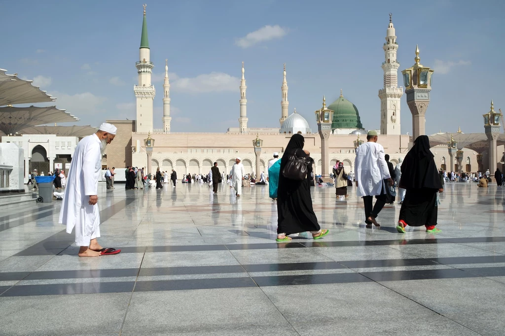 Medyna, KSA Ludzie w tradycyjnych muzułmańskich strojach spacerują po marmurowym placu przy dużym meczecie z charakterystycznymi minaretami i zieloną kopułą, otoczeni spokojną atmosferą miejsca pielgrzymkowego.