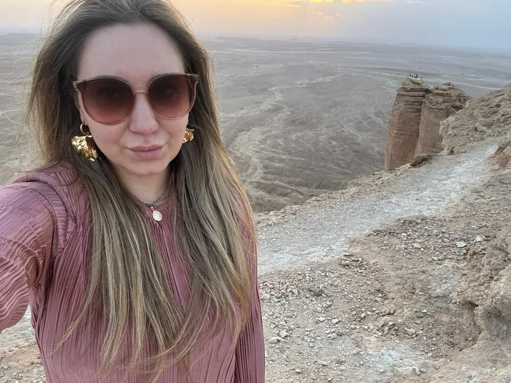 Karolina Wachowicz Kobieta w okularach przeciwsłonecznych i bluzce w odcieniu różowym robi selfie na tle pustynnego krajobrazu, widoczne są strome skały i rozległa przestrzeń, w oddali zachodzące słońce oraz jasne niebo.