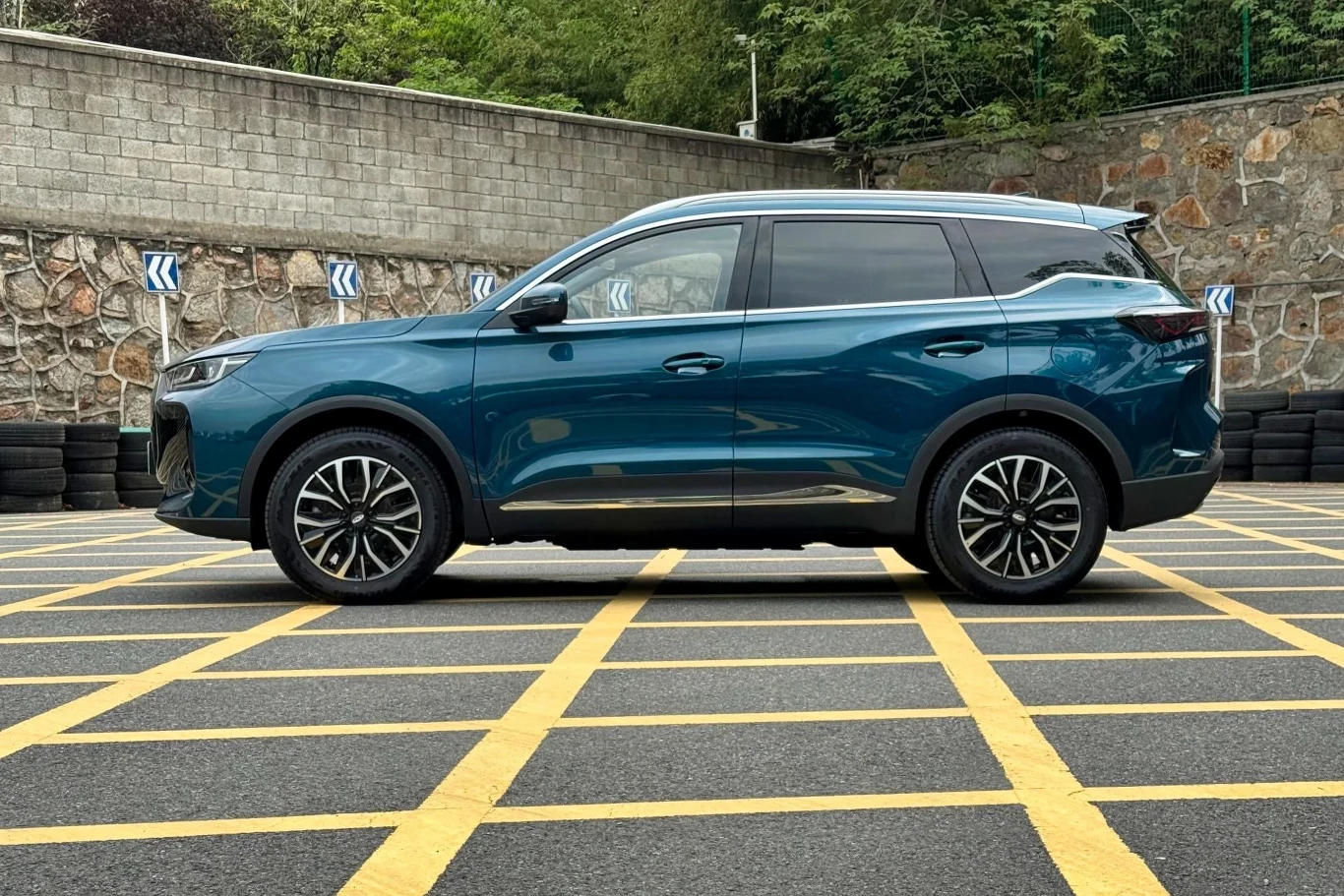 Nowoczesny, niebieski SUV zaparkowany na żółtych liniach wyznaczonego miejsca parkingowego, w tle widoczny kamienny mur i otoczenie zieleni.