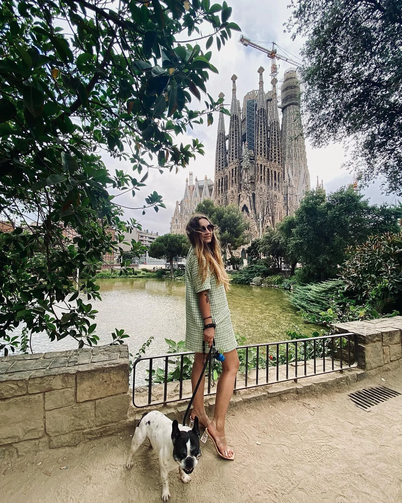 Karolina Wachowicz, w tle Sagrada Familia Kobieta w zielonej sukience i okularach przeciwsłonecznych stoi z psem na tle słynnej bazyliki Sagrada Familia w Barcelonie, otoczona zielenią i jeziorkiem