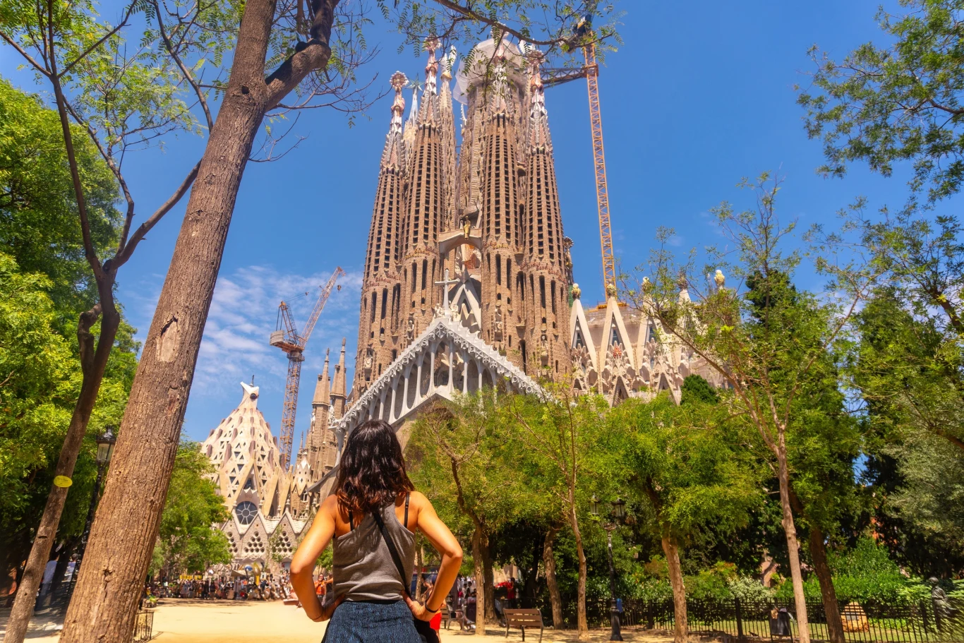 Barcelona otworzy dla zwiedzających zamknięte stacje metra Kobieta z plecakiem stoi w parku, patrząc na monumentalną bazylikę Sagrada Família, otoczoną drzewami i żurawiami budowlanymi, przy słonecznym niebie.