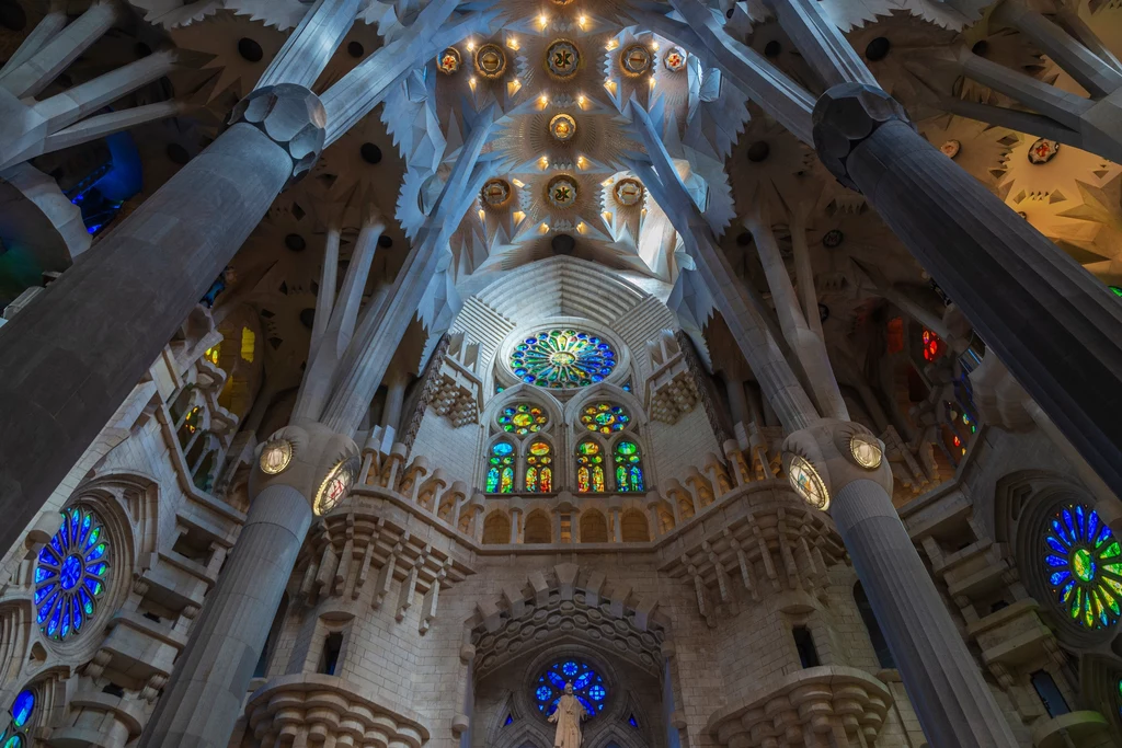 Sagrada Familia Wnętrze okazałej bazyliki z monumentalnymi kolumnami i efektownymi, kolorowymi witrażami, przez które do środka wpada wielobarwne światło, podkreślające unikalny styl architektoniczny.