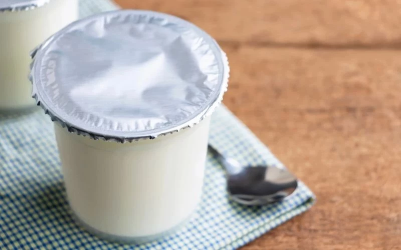 Jogurt naturalny idealnie nadaje się do marynowania drobiu Plastikowy kubek jogurtu zamknięty srebrnym wieczkiem ustawiony na błękitnej ściereczce obok łyżeczki, całość znajduje się na drewnianym stole.