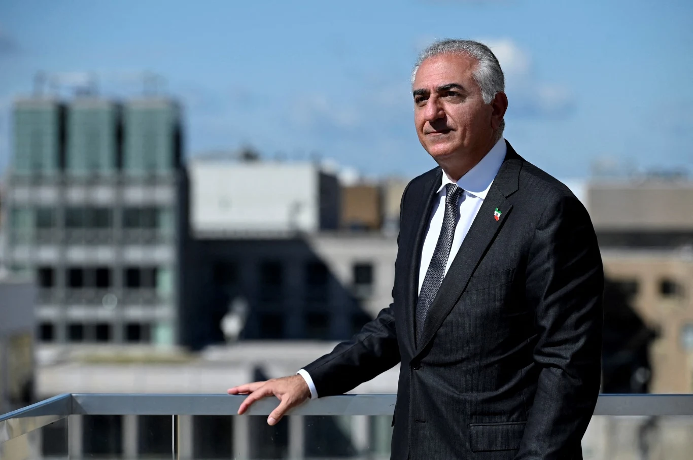 Syn ostatniego szacha Iranu Reza Pahlawi Mężczyzna w garniturze stoi na tarasie z widokiem na nowoczesne budynki miasta, trzymając jedną rękę na barierce, w tle niebo z lekkim zachmurzeniem.