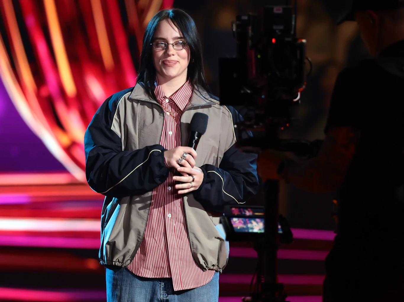 Billie Eilish Młoda kobieta z ciemnymi włosami, w okularach, trzyma mikrofon w rękach, ubrana w sportową kurtkę i koszulę w paski, stoi przed kolorowymi światłami sceny, obok znajduje się osoba z kamerą filmującą jej wystąpienie.