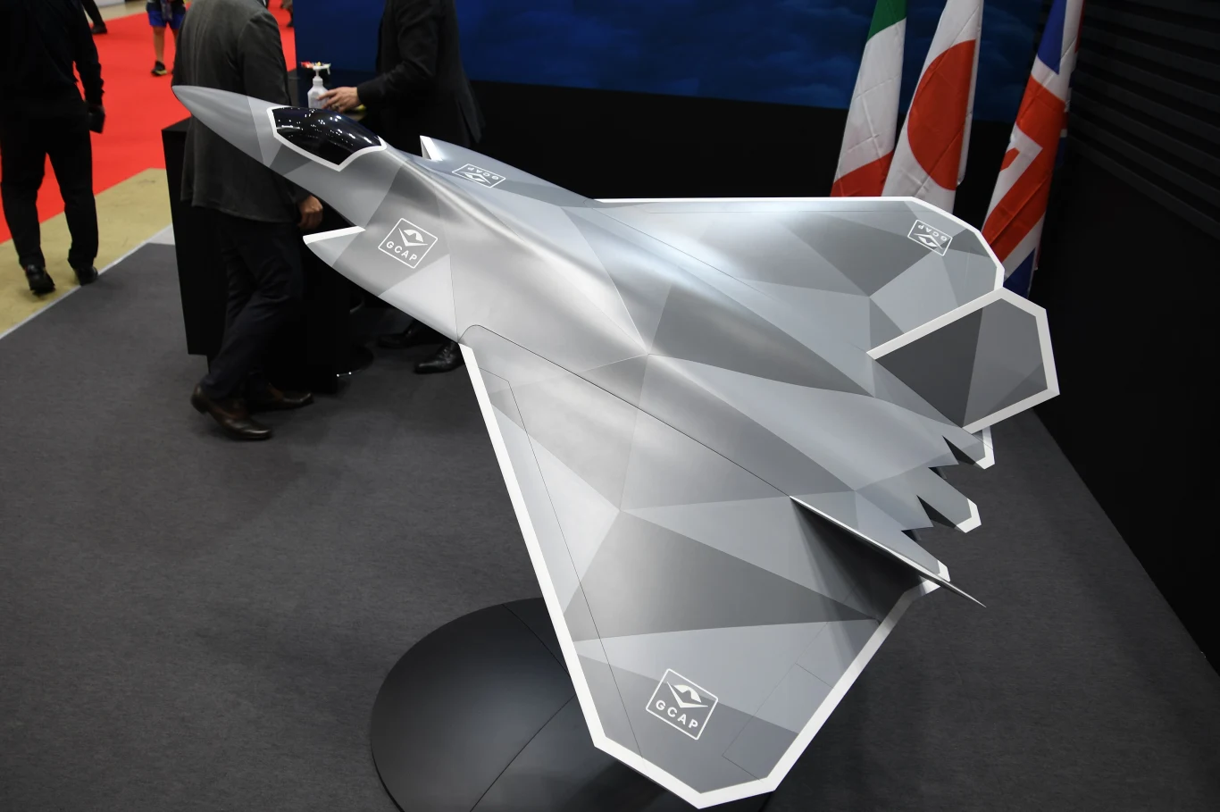 Model wizualizujący jak może wyglądać myśliwiec programu GCAP Model futurystycznego myśliwca odrzutowego o nowoczesnej, kanciastej bryle ustawiony na ekspozycji wewnętrznej, obok widoczne fragmenty flag narodowych.