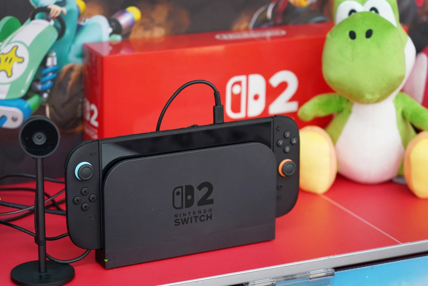 Konsola Nintendo Switch 2 w stacji dokującej przed pudełkiem z logo, po prawej stronie maskotka Yoshi z serii gier Mario, obok konsoli niewielka kamera lub mikrofon.