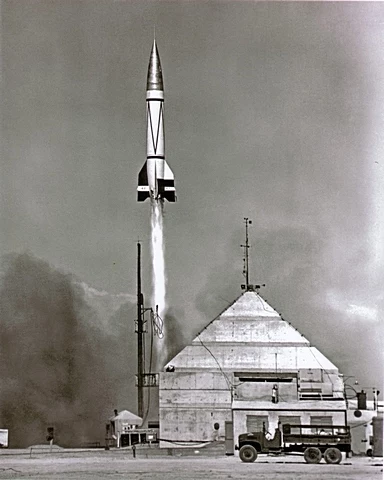 Rok 1949. Rakieta V-2 startuje w kapsułą Blossom i rezusem Albertem II na pokładzie Start rakiety z pionowej wyrzutni obok betonowego bunkra, z unoszącymi się kłębami dymu oraz widocznym w pobliżu pojazdem wojskowym, całość utrzymana w czarno-białej tonacji