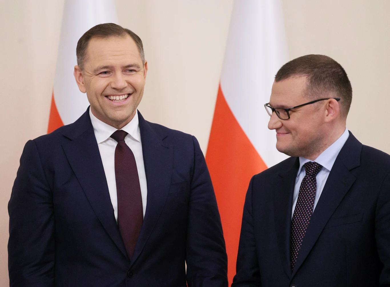 Dwóch eleganckich mężczyzn ubranych w garnitury stoi obok siebie, jeden z nich uśmiecha się szeroko, drugi patrzy w jego stronę z lekkim uśmiechem, w tle widoczne polskie flagi.