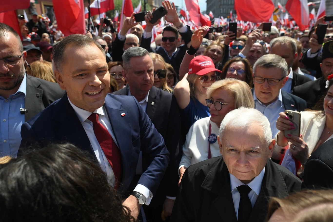 Tłum ludzi z polskimi flagami uczestniczy w wydarzeniu publicznym, na pierwszym planie znajdują się znani politycy, zza których widać uśmiechnięte twarze uczestników oraz wyciągnięte ręce wykonujące zdjęcia i gesty poparcia.