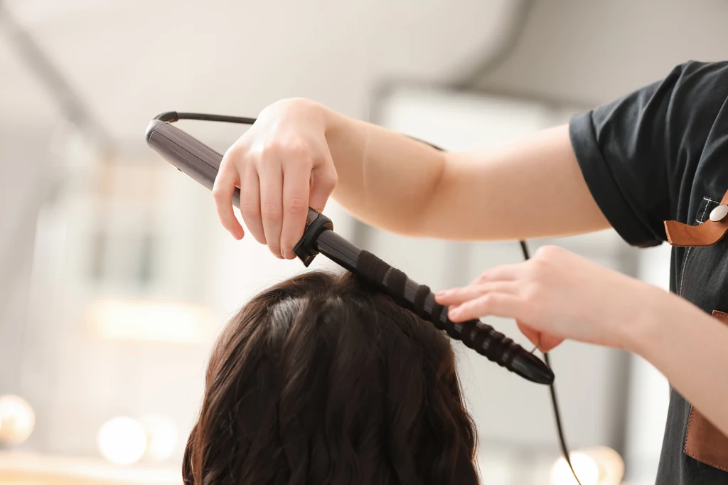 Fale są układane na włosach przy użyciu lokówki, widać ręce osoby stylizującej fryzurę.