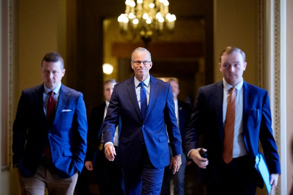 Lider senackiej większości republikanin John Thune Trzech elegancko ubranych mężczyzn idzie korytarzem, oświetlonym żyrandolem. Centralnie uchwycony mężczyzna w okularach wyróżnia się poważnym wyrazem twarzy, a po jego bokach towarzyszą mu inni dobrze ubrani mężczyźni.