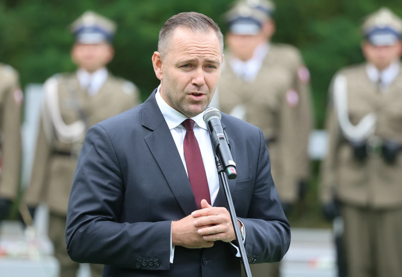 Karol Nawrocki jako prezes IPN Mężczyzna w garniturze i bordowym krawacie przemawia do mikrofonu na tle umundurowanych żołnierzy ustawionych w szeregu na zewnątrz.