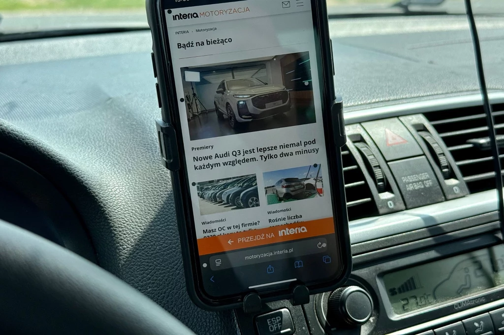 Po uruchomieniu tej funkcji, powinieneś poczuć się lepiej. Smartfon zamontowany na uchwycie samochodowym na desce rozdzielczej, na ekranie widoczny artykuł motoryzacyjny o nowym modelu Audi, otoczenie stanowią elementy kokpitu samochodu.