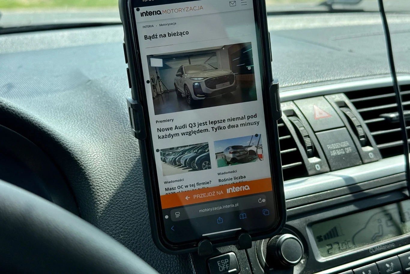 Smartfon zamontowany na uchwycie samochodowym na desce rozdzielczej, na ekranie widoczny artykuł motoryzacyjny o nowym modelu Audi, otoczenie stanowią elementy kokpitu samochodu.