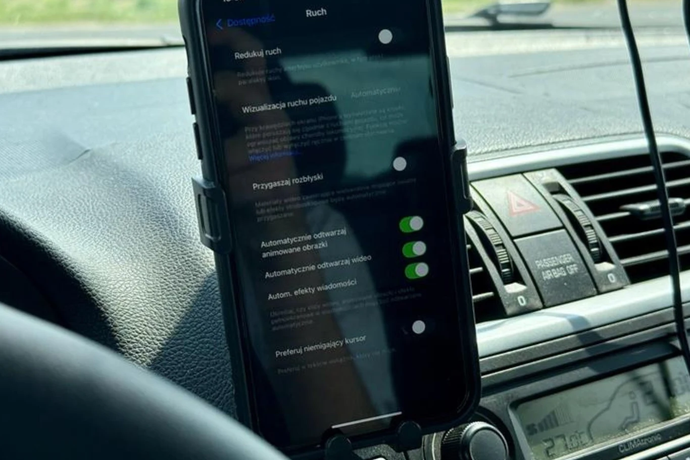 Telefon zamocowany w uchwycie na desce rozdzielczej samochodu wyświetla ustawienia systemowe, poniżej widoczne nawiewy i fragment radia, w tle rozmyty widok zewnętrzny przez przednią szybę