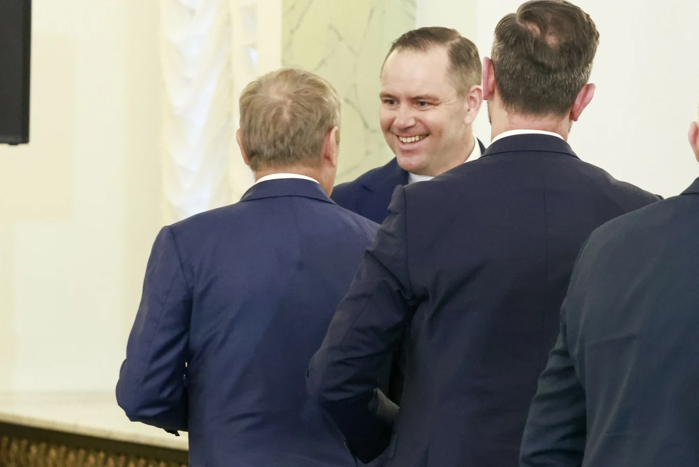 Grupa elegancko ubranych mężczyzn w garniturach prowadzących rozmowę, jeden z nich uśmiechnięty, wnętrze o jasnych ścianach z elementami ozdobnymi.