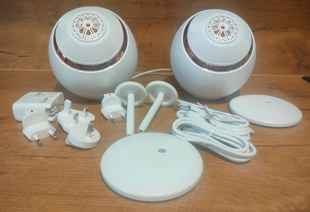 Creative Pebble Nova Zestaw dwóch białych głośników o kulistym kształcie, wraz z akcesoriami: dwie podstawki, przewody zasilające, kilka końcówek do adapterów oraz ładowarka, wszystko ułożone na drewnianej powierzchni.