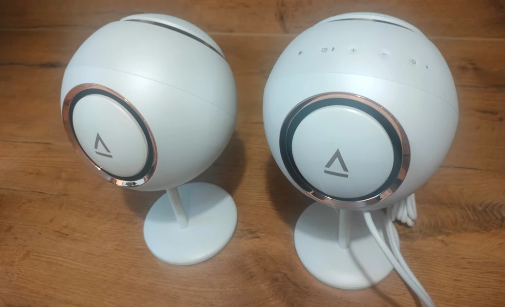 Creative Pebble Nova Dwie nowoczesne białe głośniki o kulistym kształcie stojące na płaskich podstawkach, z detalami wykończonymi metalicznymi pierścieniami, ustawione na drewnianej powierzchni.