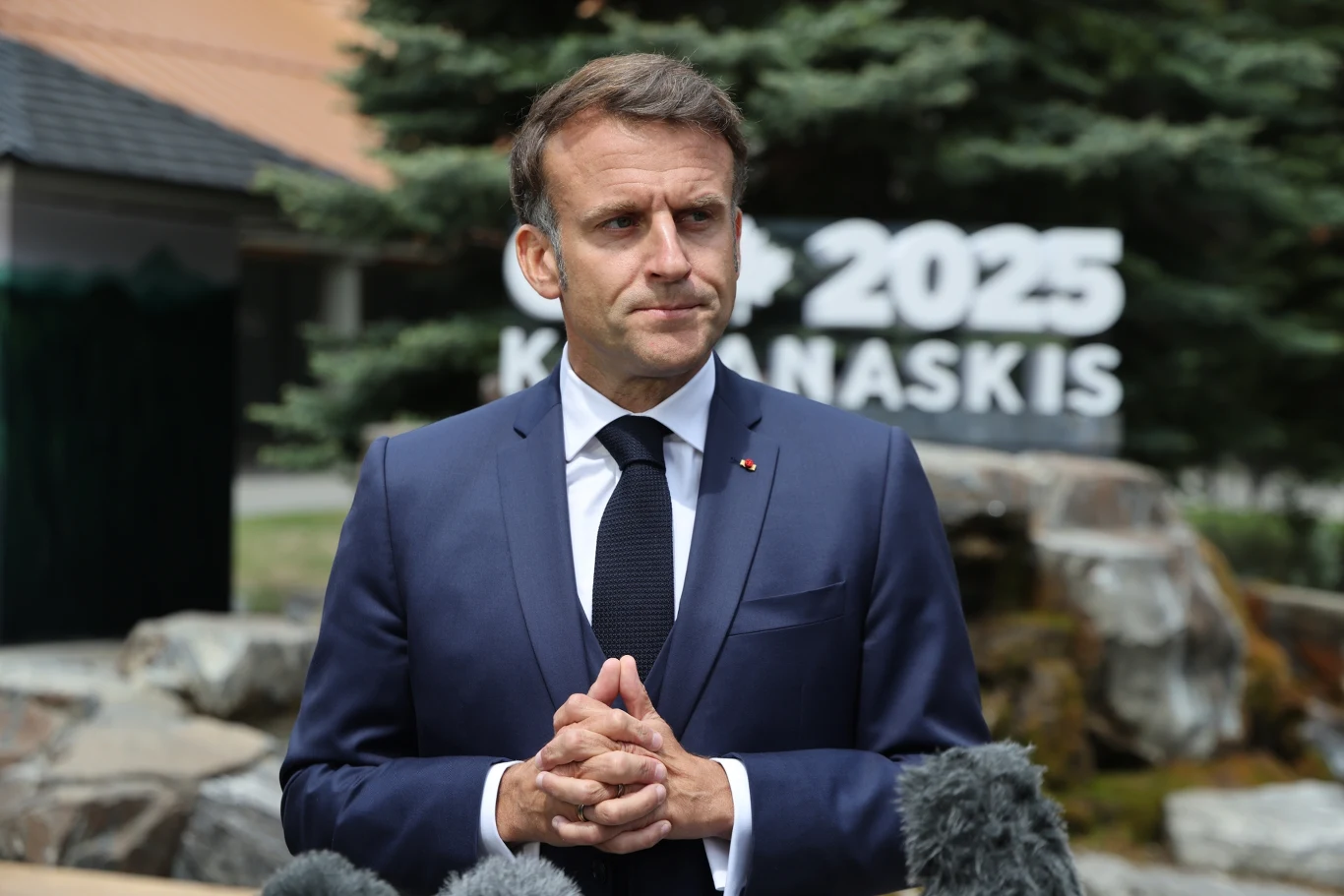 Emmanuel Macron wystąpił z inicjatywą ws. konfliktu na Bliskim Wschodzie Mężczyzna w garniturze i krawacie stoi przed kamieniami oraz tablicą z napisem G7 2025 Kananaskis, w tle zieleń, widać także kilka mikrofonów przed postacią.